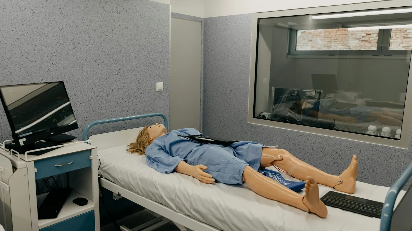 Salle de simulation d'un institut de soins, mannequin sur un lit d'hôpital à côté d'un moniteur, vitre d'observation au fond