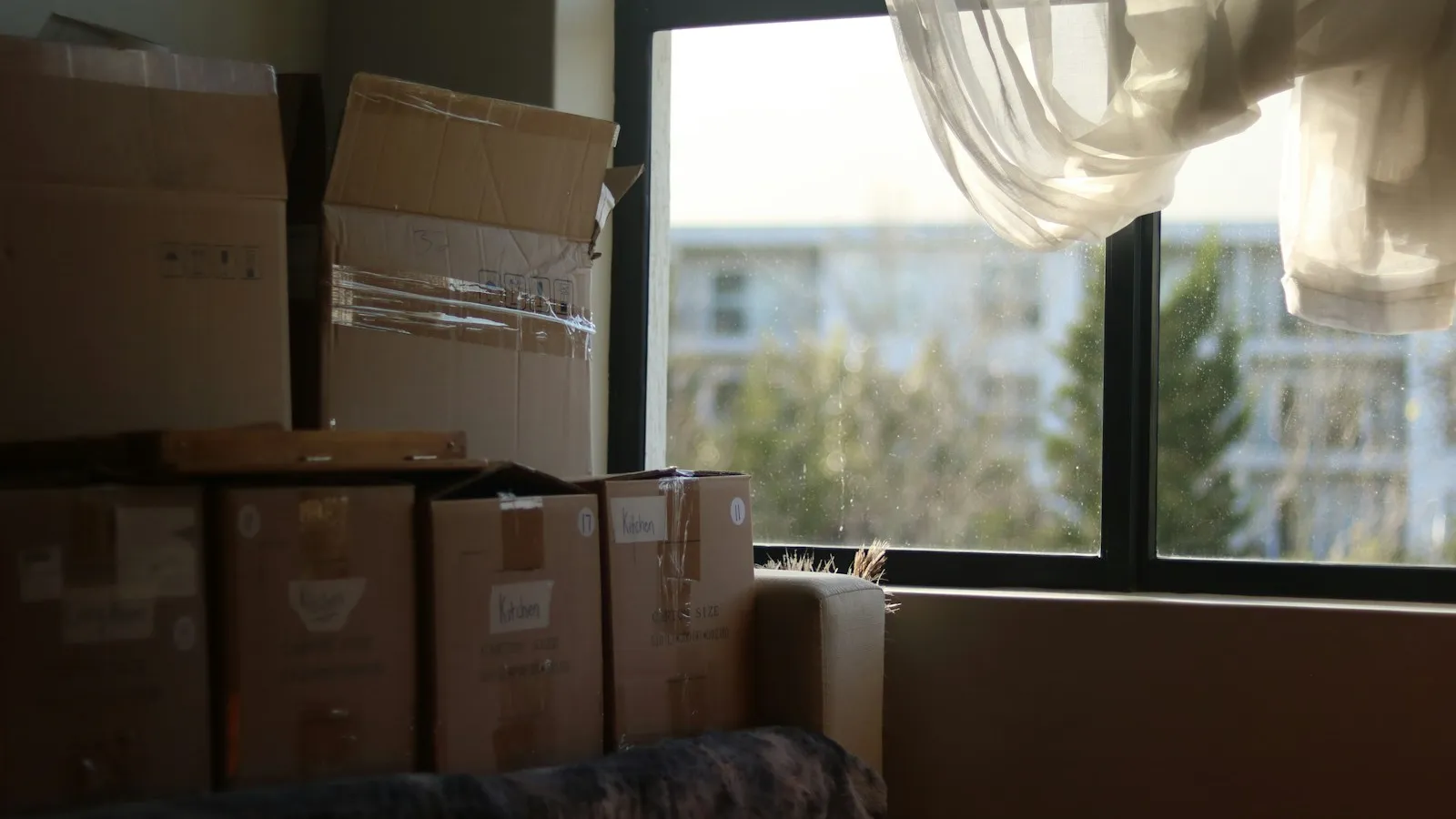 Cartons de déménagement empilés près d'une fenêtre ensoleillée, voilage blanc soulevé par le vent